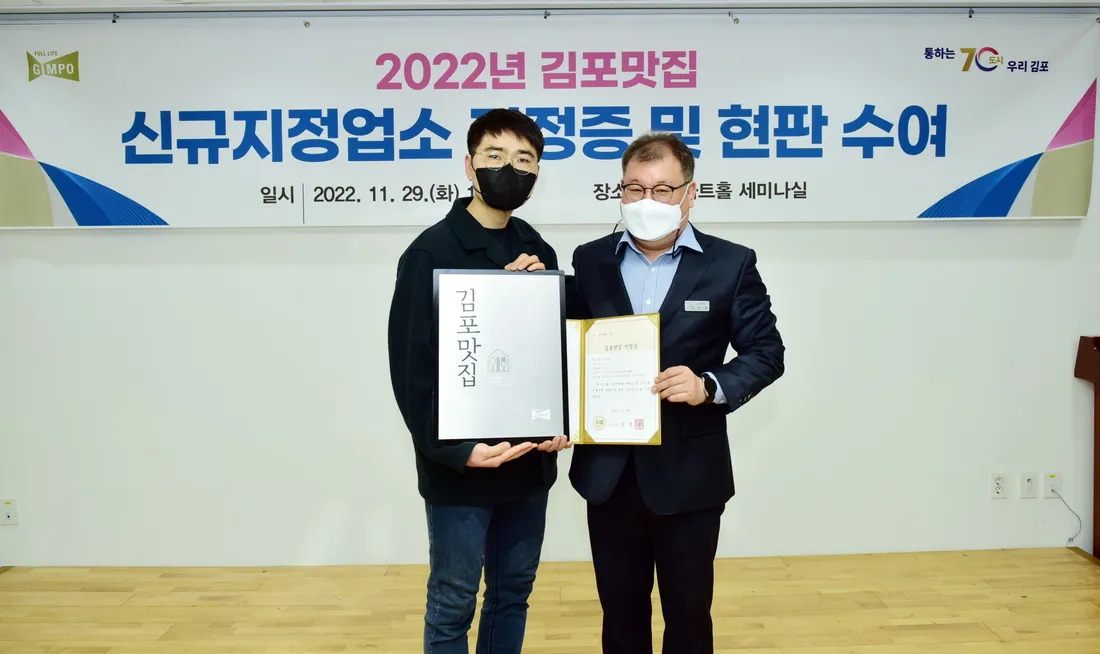 2022년 김포맛집 신규지정업소 지정증 및 현판 수여식. 산들마을 대표가 김포맛집 현판을, 김포시 관계자가 지정증을 들고 나란히 서 있다.